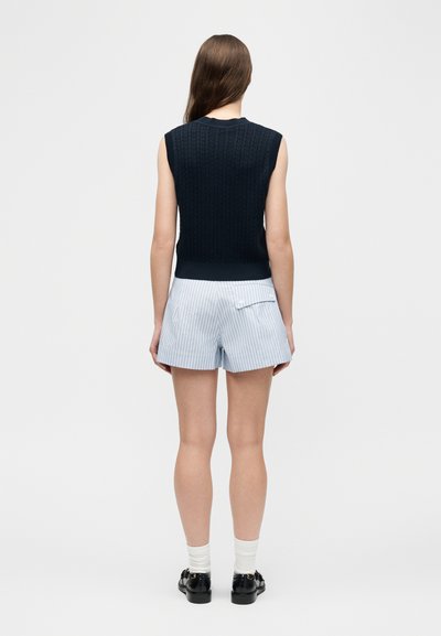 Γυναίκα με μακριά καστανά μαλλιά που στέκεται με την πλάτη, φορώντας αμάνικο μπλε πλεκτό τοπ, ανοιχτόχρωμα ριγέ σορτς, λευκές κάλτσες και μαύρα παπούτσια.