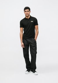 Svart poloshirt med gul logo, kombinert med svarte cargobukser. Modellen står med hendene i lommene og har på seg svarte joggesko. Enkel og moderne design.