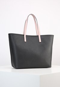 Svart tote väska med texturerad yta, med två ljusrosa axelremmar, placerad på en vit pelar mot en enkel bakgrund.