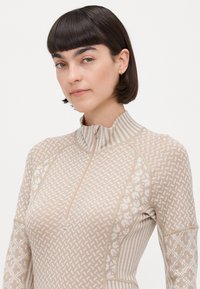 Beige hettegenser med glidelås og høy krage, med intrikate mønstre og teksturerte detaljer. Mykt, strikket materiale med ribbede detaljer.
