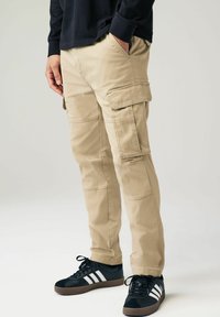 Pantaloni cargo din bumbac bej, cu buzunare laterale, tăiere dreaptă și cusături întărite, asortați cu adidași negri cu dungi albe.