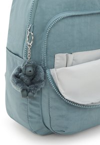 Zaino teal in tessuto con una tasca con zip e un portachiavi a forma di gorilla grigio peloso. Texture liscia e design semplice.