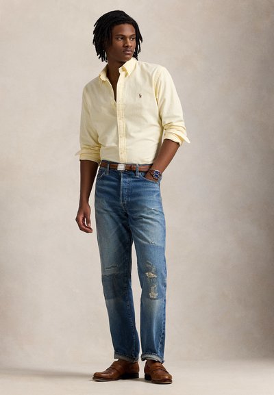 Φωτεινό κίτρινο πουκάμισο με κουμπιά, μπλε τζιν με φθορές και μπαλώματα, καφέ ζώνη και καφέ μοκασίνια· το μοντέλο στέκεται με χαλαρωμένα χέρια.