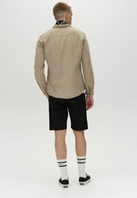 Beige Langarmhemd mit Knopfmanschetten, kombiniert mit schwarzen Shorts. Auffällige schwarz-weiße gestreifte Socken und dunkle Schuhe runden den Look ab.