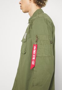 Green military-style shirt with a front patch pocket, side pockets, and a zipper pocket. Features a red ribbon label reading "REMOVE BEFORE FLIGHT."