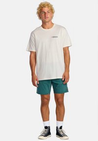 Bílé tričko s krátkým rukávem a malým logem, kombinované s tyrkysovými šortkami a černými vysokými teniskami, stojící na neutrálním pozadí.