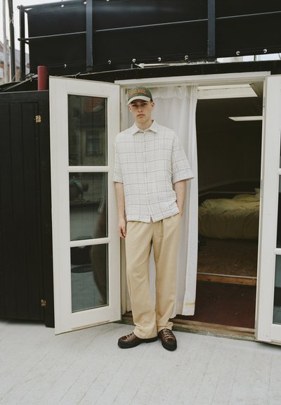Chemise à manches courtes à carreaux beige clair, associée à un pantalon beige. Chaussures foncées avec lacets. Le modèle se tient dans une porte, éclairage intérieur doux.