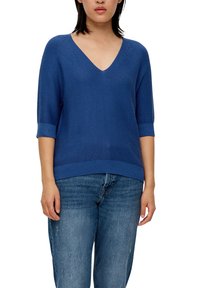 Blauer V-Ausschnitt-Pullover mit kurzen Ärmeln, gerippten Bündchen und Saum, der ein strukturiertes Strickmuster aufweist. Kombiniert mit hellblauen Jeans.