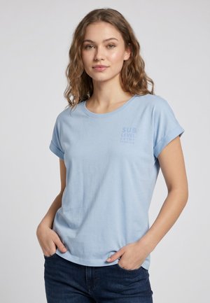 Junge Frau mit welligem hellbraunem Haar, die ein hellblaues T-Shirt mit hochgekrempelten Ärmeln und dunkelblaue Jeans trägt, mit den Händen in den Taschen.