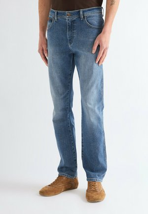 Man som bär ljusblå jeans med raka ben och bruna mockaskor med snörning, står mot en enfärgad bakgrund.