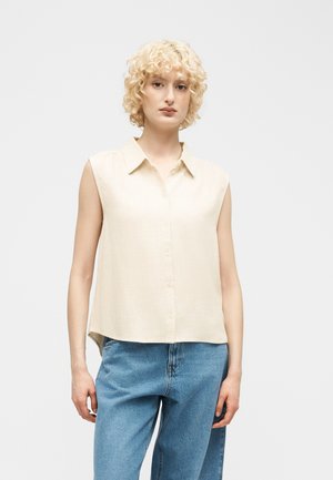 Blondínka s kučeravými vlasmi, oblečená v béžovej bezrukávovej košeli na gombíky a modrých džínsoch, stojaca na jednoduchom bielom pozadí.