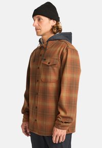 Camisa de cuadros en tonos naranjas y marrones con capucha gris, que cuenta con un cierre de botones en el frente, un bolsillo en el pecho y cordones ajustables.