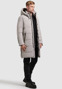 Cappotto lungo grigio imbottito con cappuccio, dotato di tasche laterali, chiusura frontale con zip e taglio dritto. Indossato sopra una maglietta nera e jeans scuri.