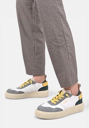 Persoon draagt grijze en witte sneakers met gele veters en accenten, gecombineerd met bruine geruite broekjes die boven de enkel zijn geknipt.