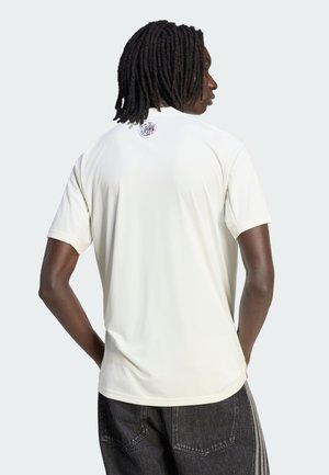 Hombre con dreadlocks que lleva una camiseta blanca de manga corta lisa y vaqueros negros, visto desde atrás sobre un fondo claro y liso.