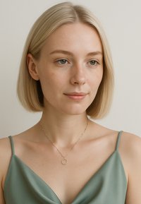 Guldkæde med en cirkulær vedhæng, prydet med små perler langs en delikat kæde. Modellen bærer en grøn satin top med tynde stropper.