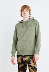 Felpa verde oliva con tasca a marsupio frontale e cappuccio con coulisse. Presenta un piccolo patch con il marchio sul petto. Abbinata a pantaloni in camouflage.