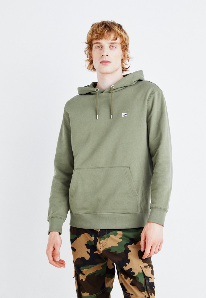 Felpa verde oliva con tasca a marsupio frontale e cappuccio con coulisse. Presenta un piccolo patch con il marchio sul petto. Abbinata a pantaloni in camouflage.