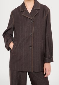 Veste marron foncé à fines rayures, avec trois boutons noirs, poches avant et coutures jaunes visibles, portée avec un pantalon assorti.