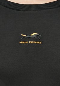 Černá mikina se žebrovaným výstřihem, na které je vyšitý logo skákajícího zvířete v černé a žluté barvě, text zní "ARMANI EXCHANGE."