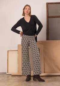 Schwarzes langärmeliges Oberteil mit V-Ausschnitt und Drehdetail, kombiniert mit cremefarbenen und schwarzen, gemusterten, weiten Hosen. Modell trägt dunkle Stiefeletten.