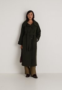 Casaco longo verde-oliva com um colarinho largo, cintura cingida e dois botões. O modelo usa calças castanhas e segura uma mala bordô.