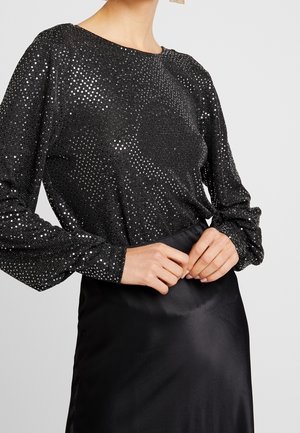 Vrouw die een zwart shirt met lange mouwen en een zilverkleurig sprankelpatroon draagt, met een zwarte satijnen rok, handen in elkaar gevouwen ter hoogte van de taille.