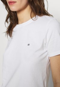 Femme portant un t-shirt blanc à manches courtes avec un petit logo noir "ck" sur la poitrine et des cheveux bruns lâchés visibles.