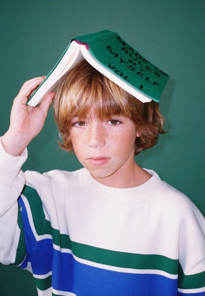 Un bambino tiene un libro verde con testo nero sulla testa. Indossa un maglione bianco con strisce blu e verdi. Lo sfondo è di un verde solido.