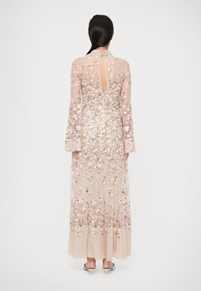 Γυναίκα που φοράει ένα μακρύ διάφανο μπεζ φόρεμα με ροζ και πράσινα λουλούδια κεντημένα, με άνοιγμα στην πλάτη και μακριά μανίκια, στέκεται μπροστά σε έναν απλό λευκό φόντο.