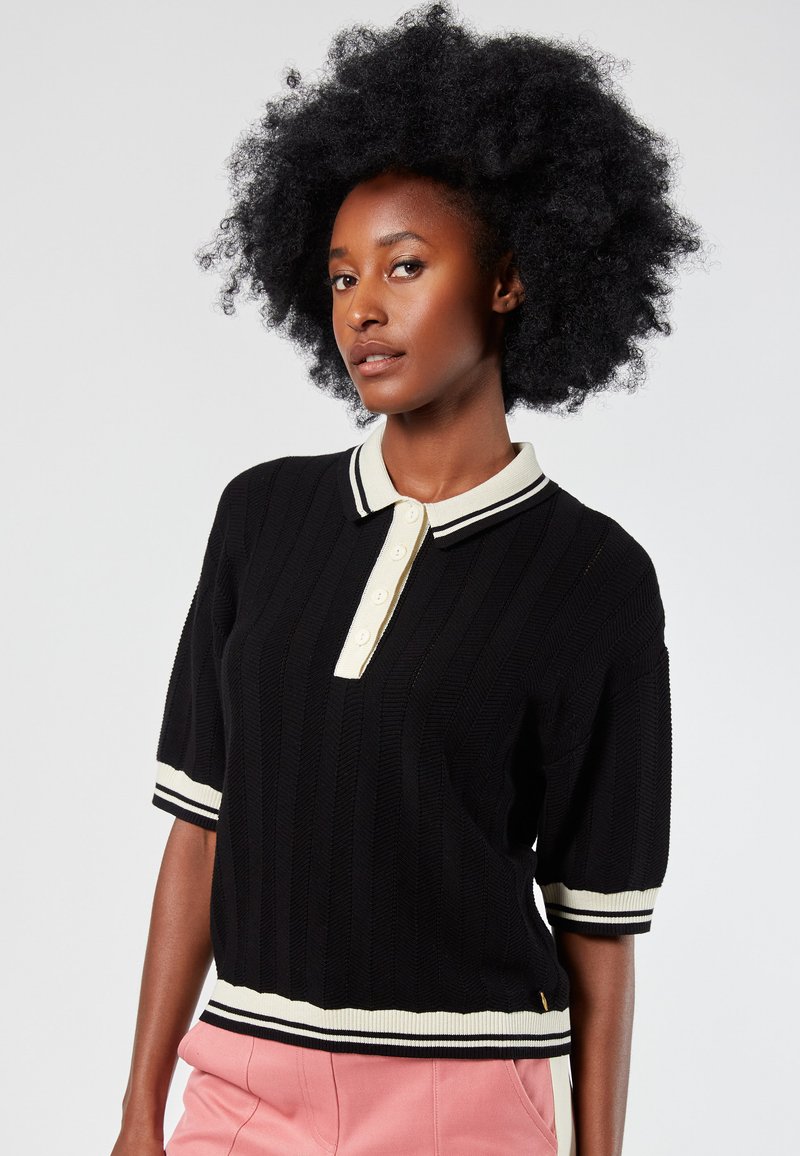 Camisa polo negra con rayas en relieve y cuello crema, con tapeta de cuatro botones y acentos a rayas en las mangas y el dobladillo. Mangas cortas, tejido texturizado.