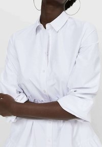 Blusa blanca de botón con un cuello estructurado, cintura ajustada y mangas de tres cuartos, que presenta una textura de algodón suave y nítida.