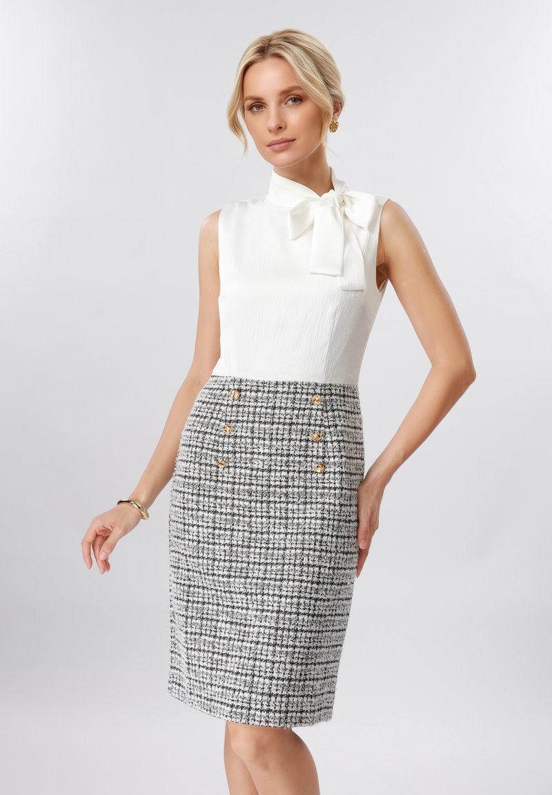 Femme portant un chemisier blanc sans manches avec un nœud et une jupe à carreaux noir et blanc taille haute avec des boutons dorés, posant devant un fond uni.