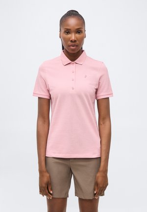 Mujer de piel oscura y cabello recogido, vestida con una camiseta polo de manga corta de color rosa claro y pantalones cortos beige hasta la rodilla, de pie frente a un fondo blanco.