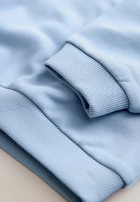 Tejido de sudadera azul claro con puños y dobladillo acanalados. Textura suave con ligero estiramiento, doblado para mostrar capas y costuras.