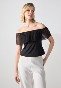 Top negru, cu umerii expuși, decorat cu un overlay din voal cu volane, realizat dintr-un material neted, asortat cu pantaloni albi cu picioare largi.