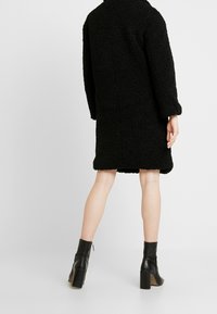 Černý fleecový kabát s texturovaným, rastrovaným povrchem, se rovnoměrným střihem, dlouhými rukávy a délky po střed stehen, doplněný černými kotníkovými botami.