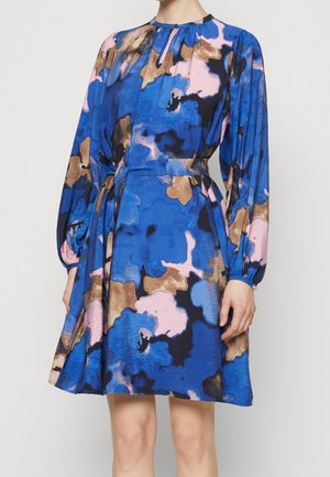 Vrouw die een knie-lange jurk draagt met lange pofmouwen en een abstract patroon in blauw, roze, zwart en bruin, voorzien van een ceintuur in de taille.