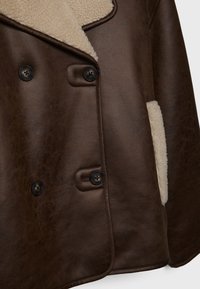 Manteau marron à double boutonnage avec des boutons noirs et une doublure en shearling crème visible au niveau du col et du bord des poches.