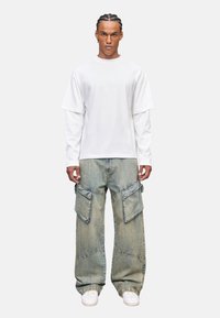 Weißes kurzärmeliges, geschichtetes T-Shirt kombiniert mit hellblauen, weiten Cargo-Jeans mit großen Taschen und strukturierter Oberfläche, getragen mit weißen Sneakers.