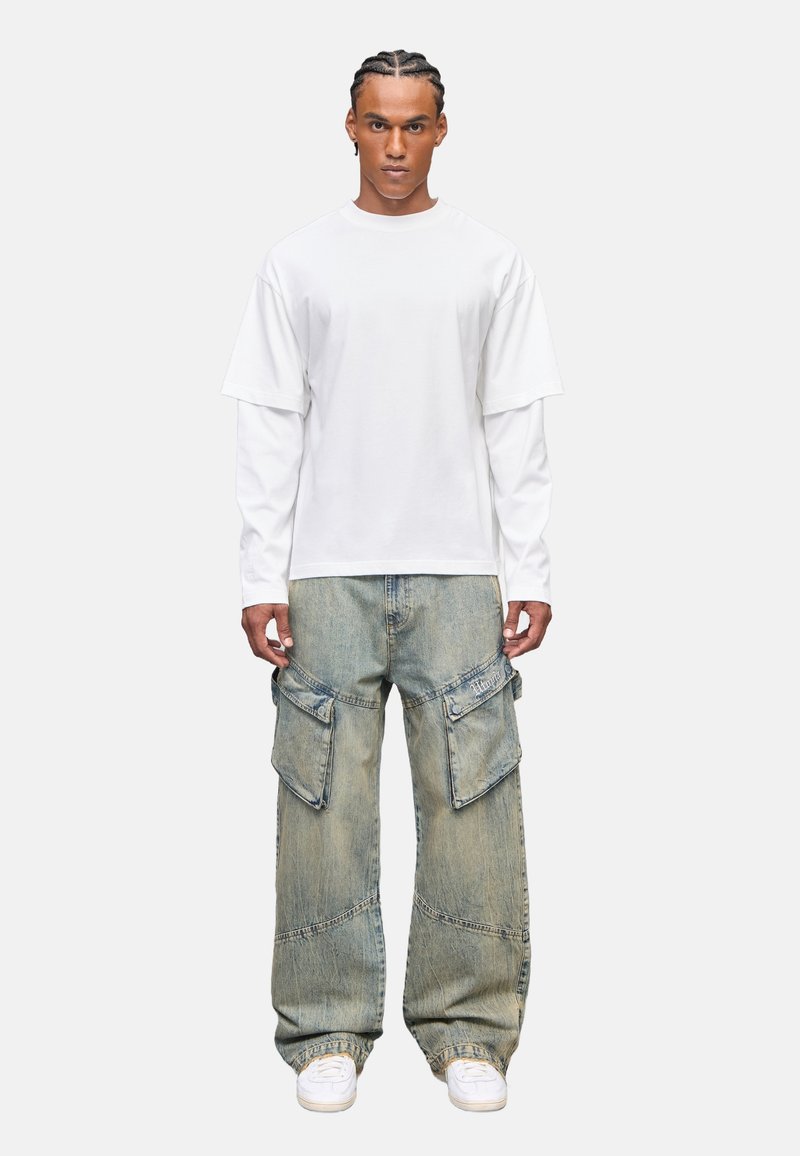 Weißes kurzärmeliges, geschichtetes T-Shirt kombiniert mit hellblauen, weiten Cargo-Jeans mit großen Taschen und strukturierter Oberfläche, getragen mit weißen Sneakers.
