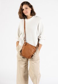 Bruine leren crossbody tas met een zachte, slungelige vorm, verstelbare schouderband en contrasterende hardwaredetails; gedragen door een model in lichte kleding.