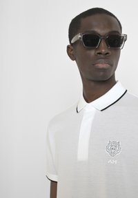 Polo blanca con ribetes negros, que presenta un logo de tigre bordado y las iniciales en el pecho, hecha de una tela suave y con un diseño ajustado.