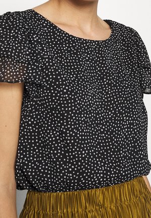 Zwarte blouse met witte polkadots en korte ruches aan de mouwen, gecombineerd met een geplooide gouden rok, getoond van nek tot taille.