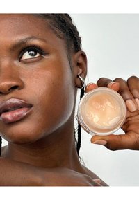 Producto de cuidado de la piel claro en un tarro transparente sostenido en una mano. El modelo tiene una piel suave y cabello trenzado, con un color de labios natural.