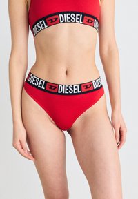 Ensemble de bikini rouge composé d'un haut et d'un bas en tissu lisse, avec une taille de marque noire ornée de lettres blanches.