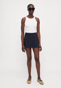 White sleeveless tank top paired with a navy pleated skirt. Model wears black cat-eye sunglasses and tan slip-on shoes. Simple and fitted design.