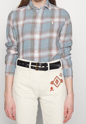 Personne portant une chemise à carreaux bleu et marron avec des boutons, un pantalon blanc avec une broderie orange sur la poche, et une ceinture noire à double boucle.