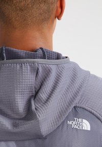 Grå hoodie i ett texturerat tyg, med stor huva och en logotyp på baksidan. Materialet verkar vara lätt och andningsbart.