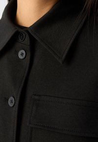 Czarna koszula z klasycznym kołnierzykiem, wyposażona w dwa czarne guziki i kieszeń na piersi. Gładka tekstura z delikatnym przeszyciem.